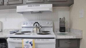Cozinha com fogão branco, chaleira e escorredor de louça. Há armários acima do fogão e um porta-facas na bancada.