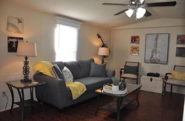 A living room with a grey sofa, a small table with a lamp, a ceiling fan, and various wall decorations including framed pictures and a guitar. - Low Rent Rendezvous (Lebanon)