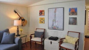 Sala de estar com um violão, duas poltronas, uma mesa com abajur e obras de arte nas paredes.