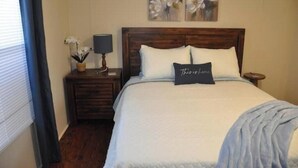Quarto com cabeceira de madeira, colcha branca, travesseiro com a inscrição 'This is home' e pintura de flores na parede.