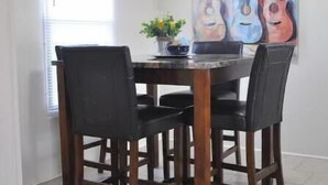 Uma sala de jantar com uma mesa de madeira e quatro cadeiras, um quadro de guitarras na parede e uma janela com persianas.