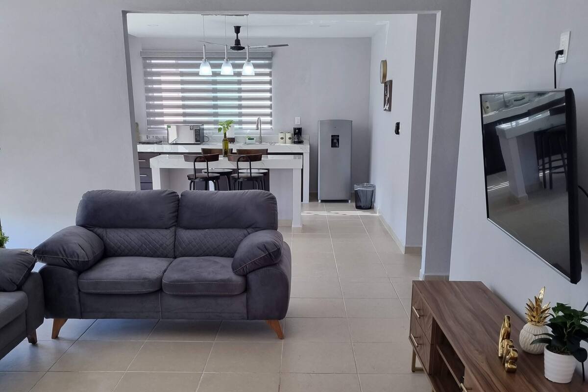 A modern living room with a grey sofa, a wooden TV stand, and a kitchen in the background.
