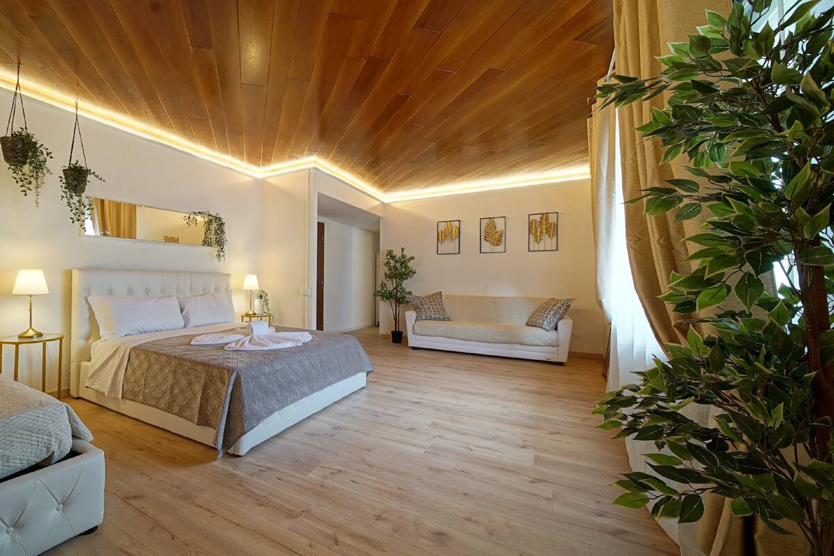 A hotel room with a wooden ceiling, two beds, a sofa, and several potted plants.