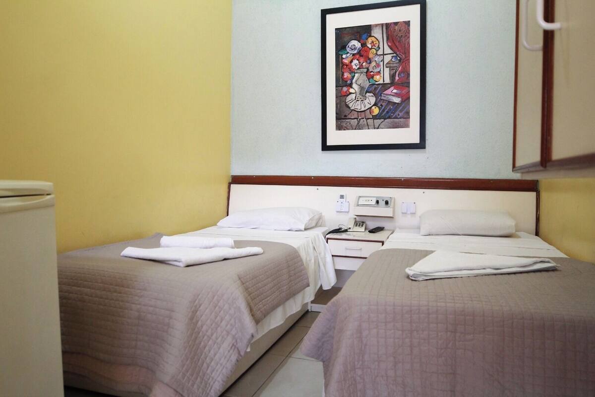 Hotelkamer met twee bedden, een nachtkastje met telefoon, een klein schilderij aan de muur en een raam met gordijnen.
