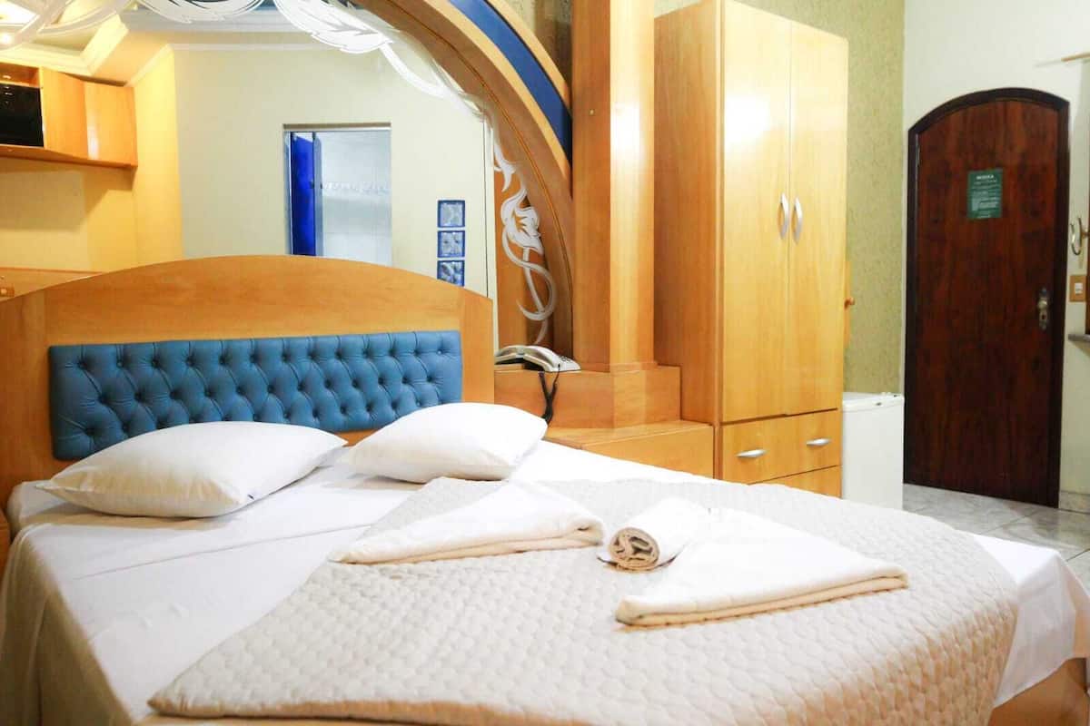 Een netjes opgemaakt bed met een blauw hoofdbord, een houten garderobe en een deur met een bordje.