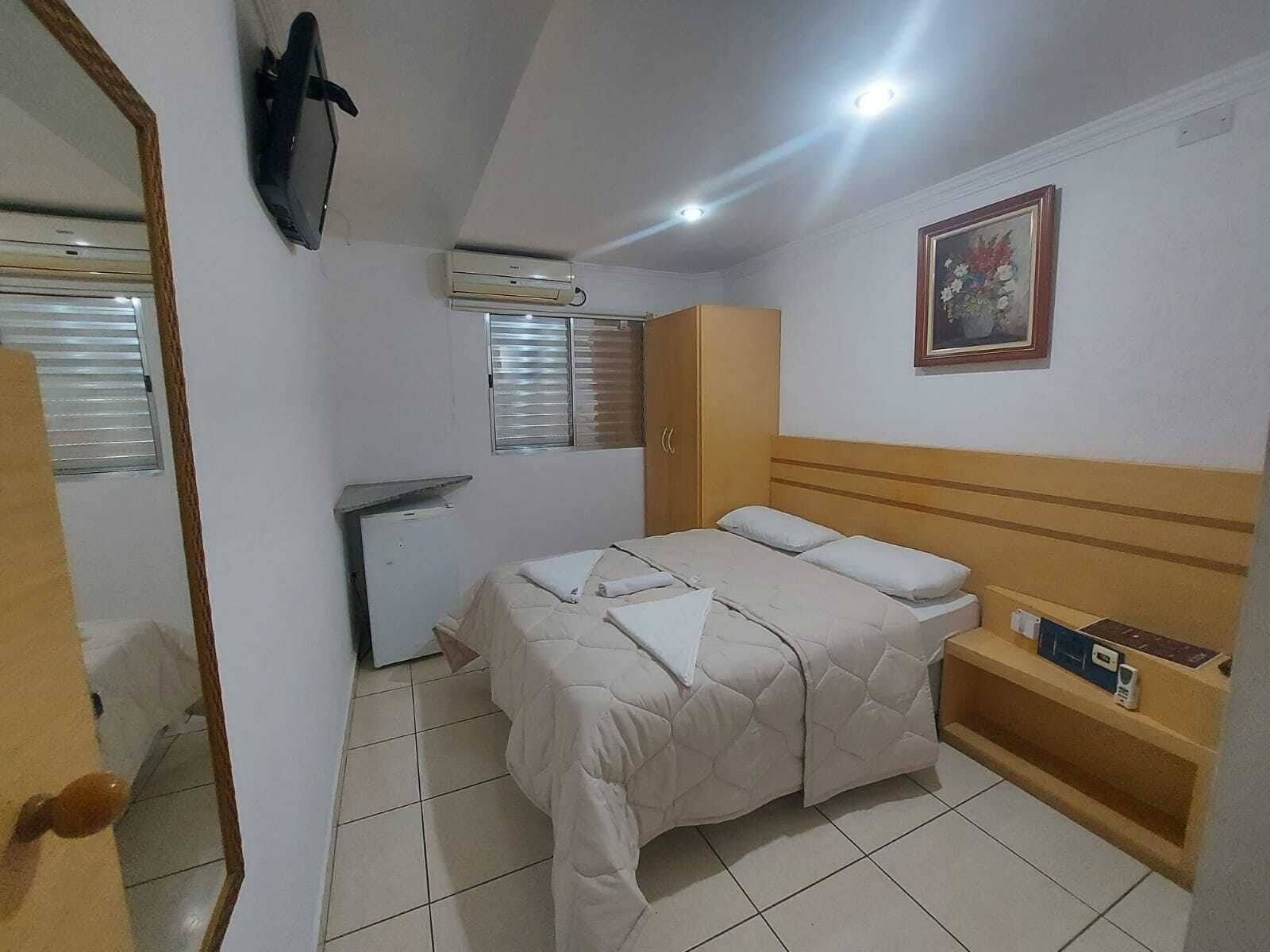 Um quarto pequeno e limpo, com uma cama, uma escrivaninha, uma cadeira, uma televisão e uma janela.