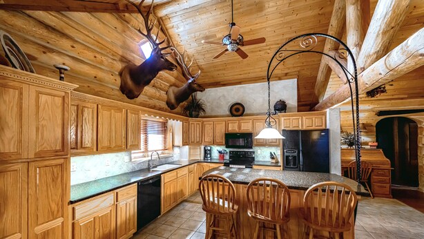 A spacious kitchen with wooden cabinets, a central island with bar stools, a ceiling fan, and a mounted deer head. - Secluded & Spacious Log Cabin Lodge Located in Western Nebraska - Pet-friendly! (Lodgepole)