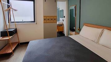 Una camera d'albergo con un letto, una mensola in legno, un appendiabiti, una finestra, un bagno con uno specchio e una parete decorata.
