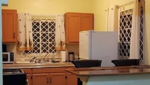 Cocina compacta con armarios de madera, microondas y refrigerador. Hay una zona de comedor con una silla a rayas y una mesa pequeña.