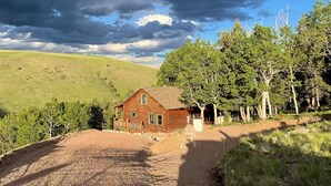 A wooden cabin with a gravel driveway, surrounded by trees and a hilly landscape. - Mountain Meadow Cabin in Cotopaxi on 40 acres (Cotopaxi)