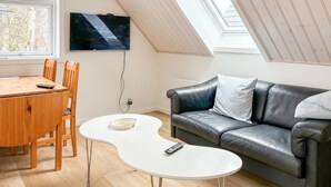 A cozy living room with a wooden dining table, a white coffee table, a black leather sofa, and a TV mounted on the wall.