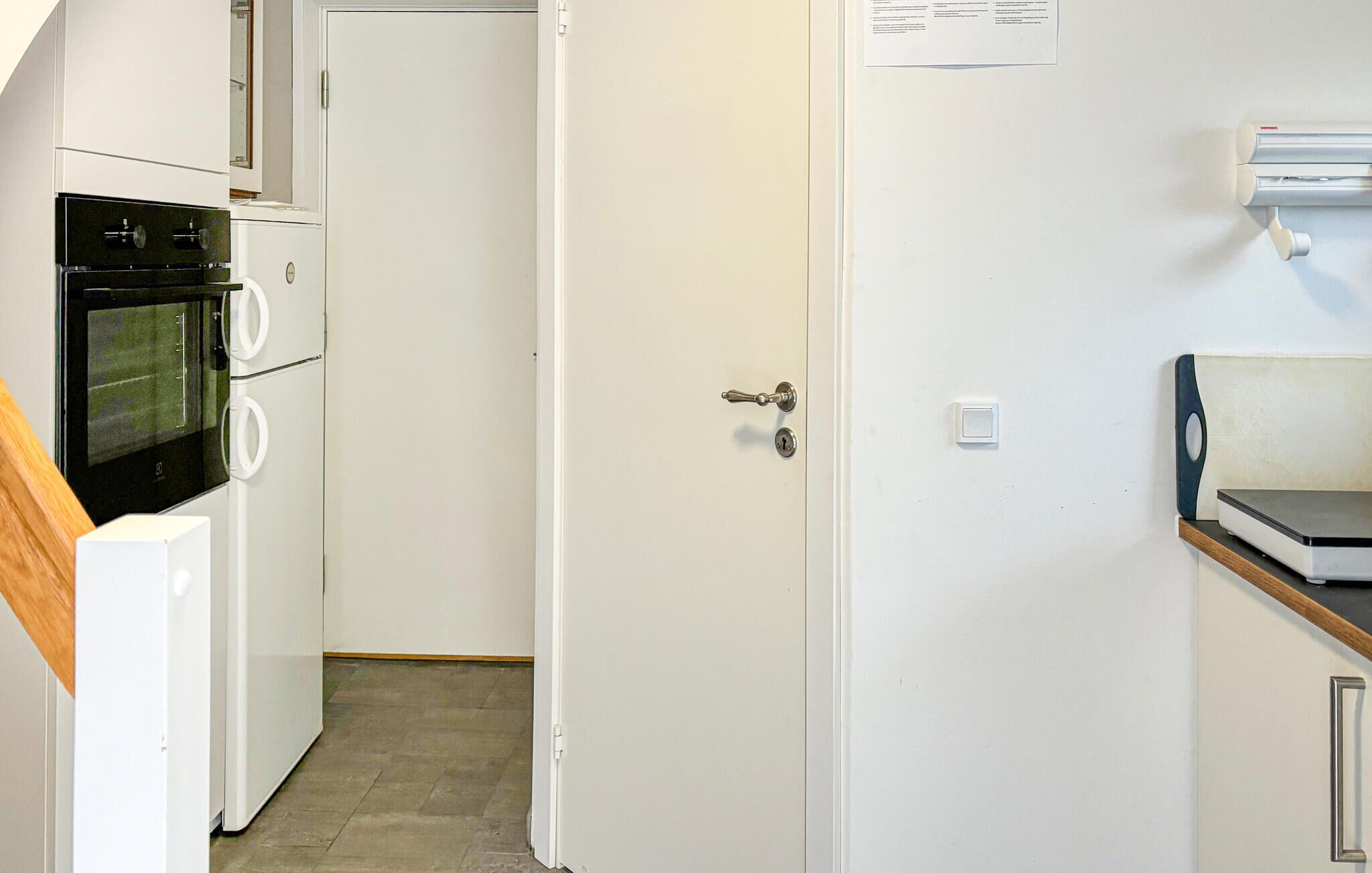A compact kitchen with a built-in oven, a microwave, and a refrigerator. A door with a key lock is visible, and a small shelf with a microwave is on the right.