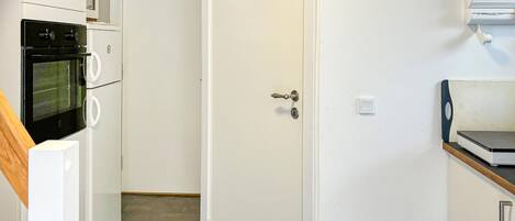 A compact kitchen with a built-in oven, a microwave, and a refrigerator. A door with a key lock is visible, and a small shelf with a microwave is on the right.