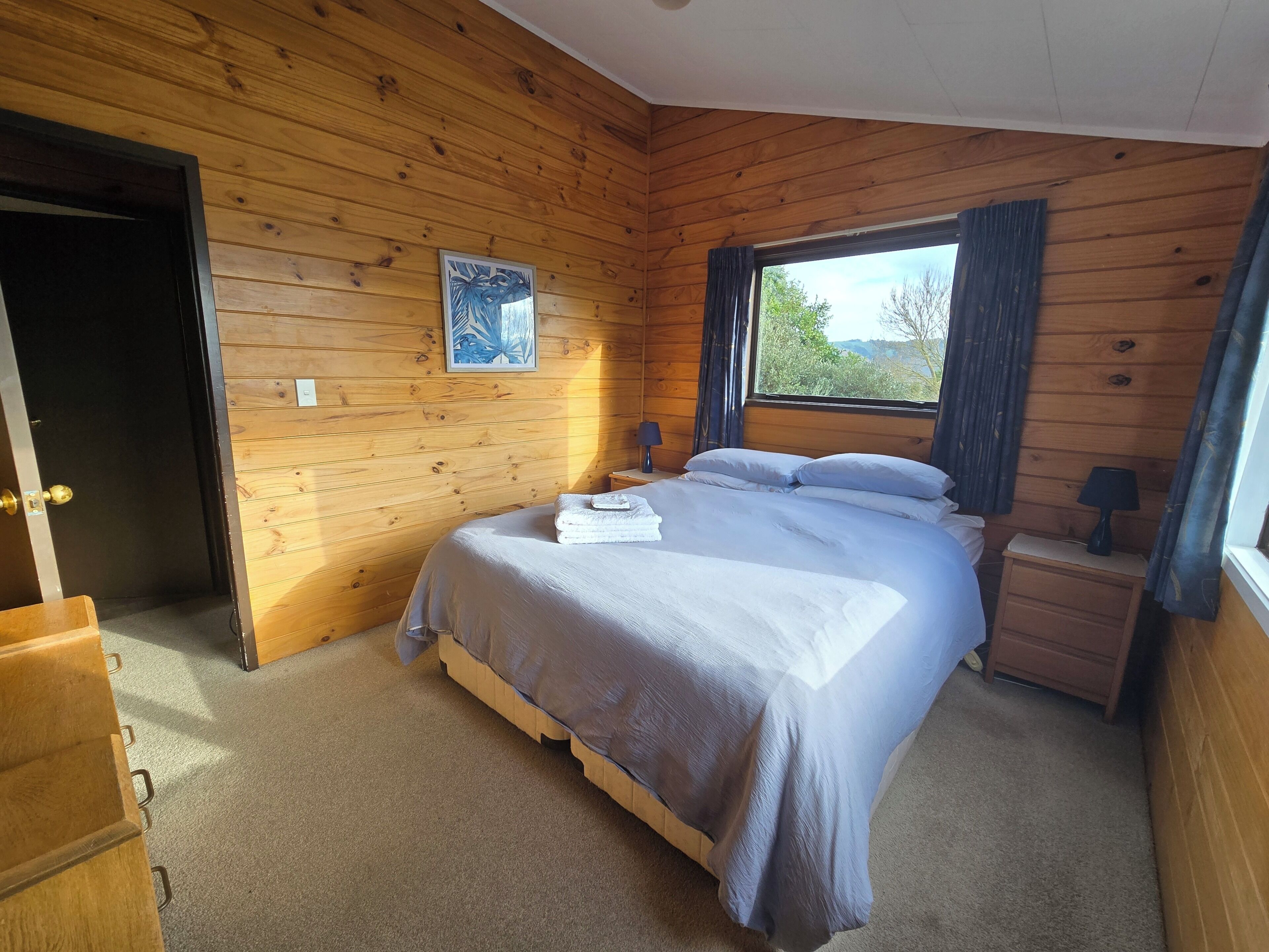 Un dormitorio con paneles de madera, una cama, una mesita de noche, una lámpara y una ventana con cortinas.