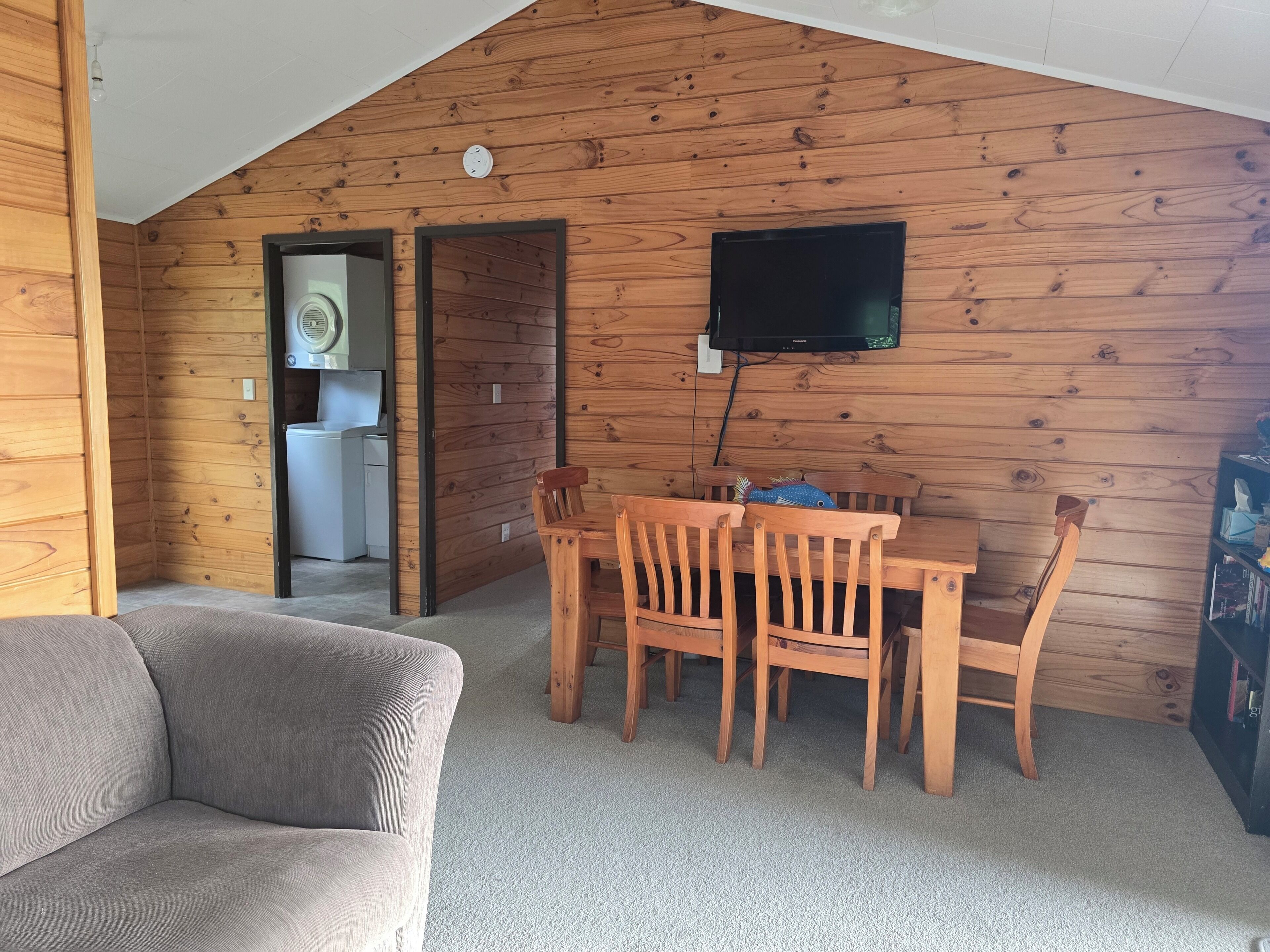 Ein gemütliches Zimmer mit Holzbalkendecke, einem Esstisch mit Stühlen, einem an der Wand montierten Flachbildfernseher und einem grauen Sofa.