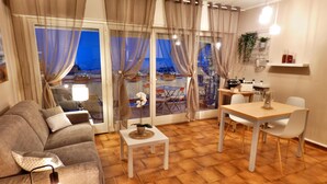 Una sala de estar moderna con sofá, mesa de comedor y sillas. Hay un ventanal con cortinas, una lámpara y una planta pequeña en un estante.