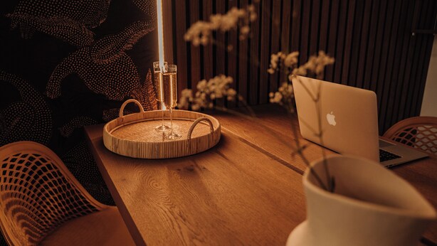 Une table en bois sur laquelle se trouve un ordinateur portable, un plateau en osier avec deux coupes de champagne et un vase décoré de branches.