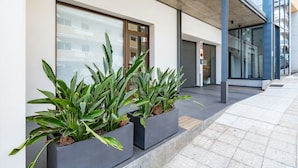Il s’agit d’un bâtiment moderne doté de grandes fenêtres et d’un auvent en béton. Sur le trottoir, on trouve deux jardinières rectangulaires contenant des plantes vertes.