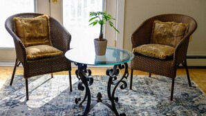 Twee rieten stoelen met bloemenkussens, een glazen tafel met een potplant en een gekleurd vloerkleed op een houten vloer.