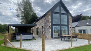 A stone building with a large glass window, a covered patio, and a fenced outdoor area. - Essich Park - 3BR - Hot Tub - Sleeps 6 (Essich)