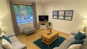 A modern living room with a flat-screen TV, grey sofas, wooden coffee table, and a view of the street through large windows.