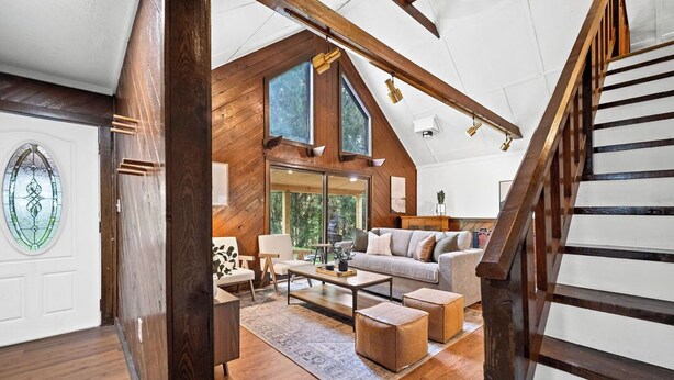 A spacious living room with a high ceiling, wooden beams, a large window, and a staircase.