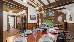 A dining area with a wooden table set for a meal, a kitchen in the background, and a view of the outdoors.
