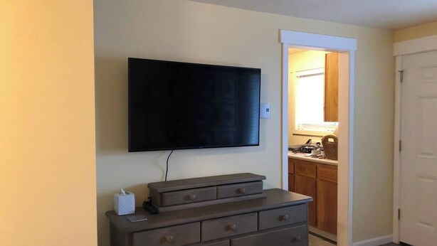 Ein Zimmer mit einem wandmontierten Flachbildfernseher, einer Holzkommode mit Schubladen und einer Tür zu einem Küchenbereich.