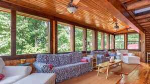 Ein geräumiges Zimmer mit Holzbalkendecke, großen Fenstern, einem gemusterten Sofa, einem weißen Sessel und einem kleinen Tisch.