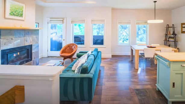 A modern kitchen with a built-in stove, a dining area with a table and chairs, a green sofa, and a view of the outdoors through large windows.