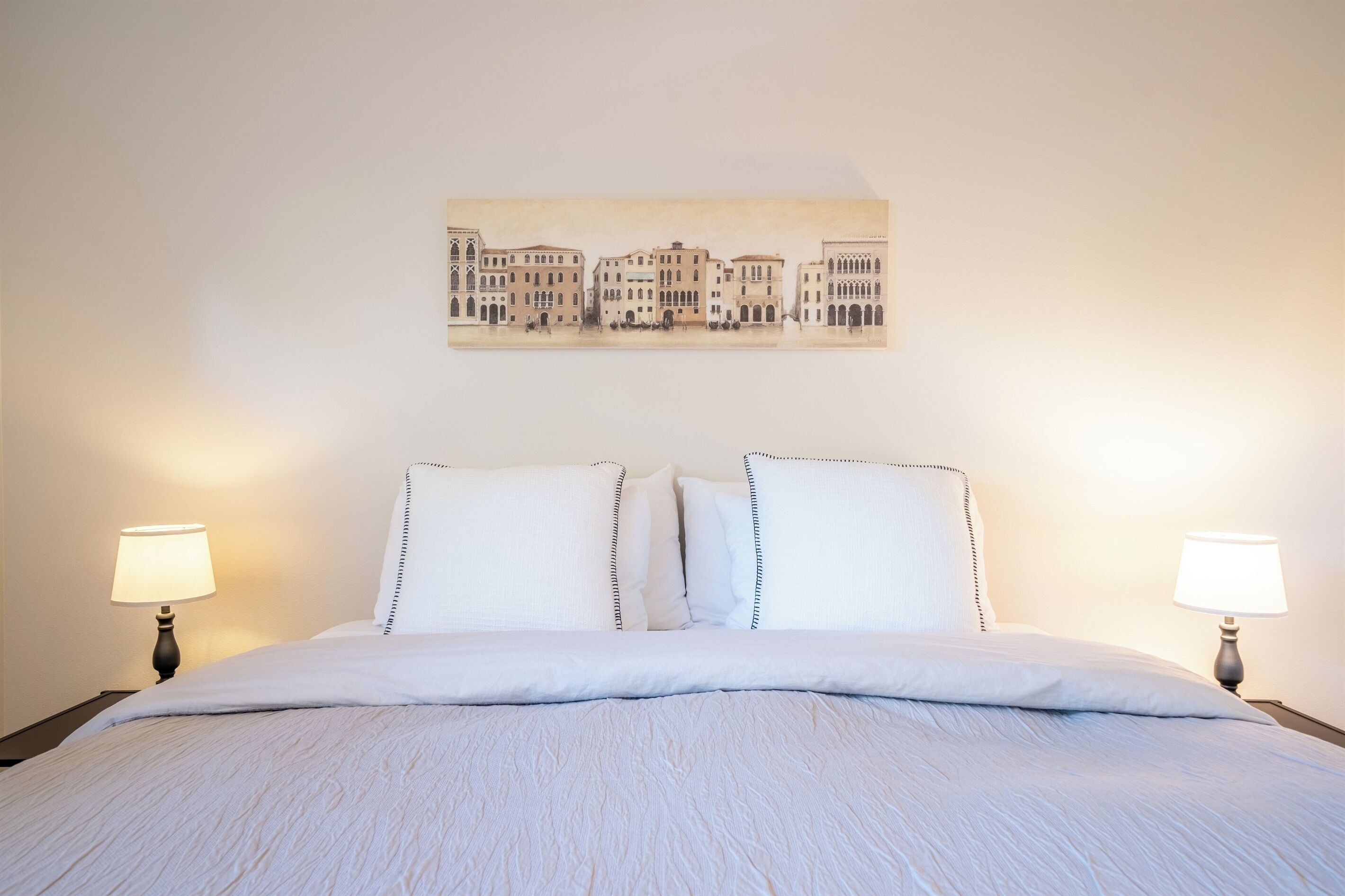 A neatly made bed with white linens and two bedside lamps, a wall-mounted artwork depicting buildings, and a simple, clean room layout.