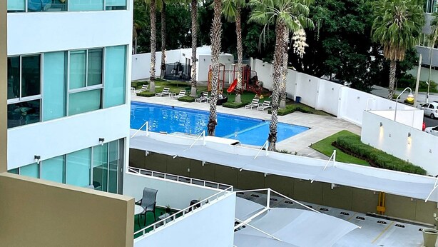 Um edifício moderno com área de piscina no terraço, rodeado por palmeiras e uma vista da linha do horizonte da cidade ao fundo.