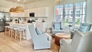 Una cocina moderna con armarios blancos, una isla central y taburetes de bar. Hay dos sillones grises y un otomano redondo de mimbre. Un candelabro cuelga del techo, y un ventanal grande proporciona luz natural.