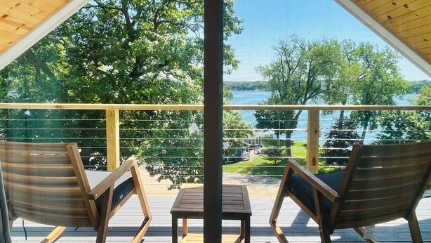 Terraza de madera con dos mecedoras y una mesa, con vista a un lago rodeado de árboles y una zona cubierta de césped.