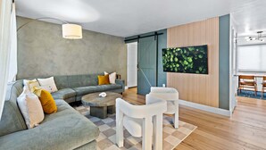 Una sala de estar moderna con un sofá gris, una mesa de centro y sillas blancas. Hay un televisor montado en la pared y piso de madera.
