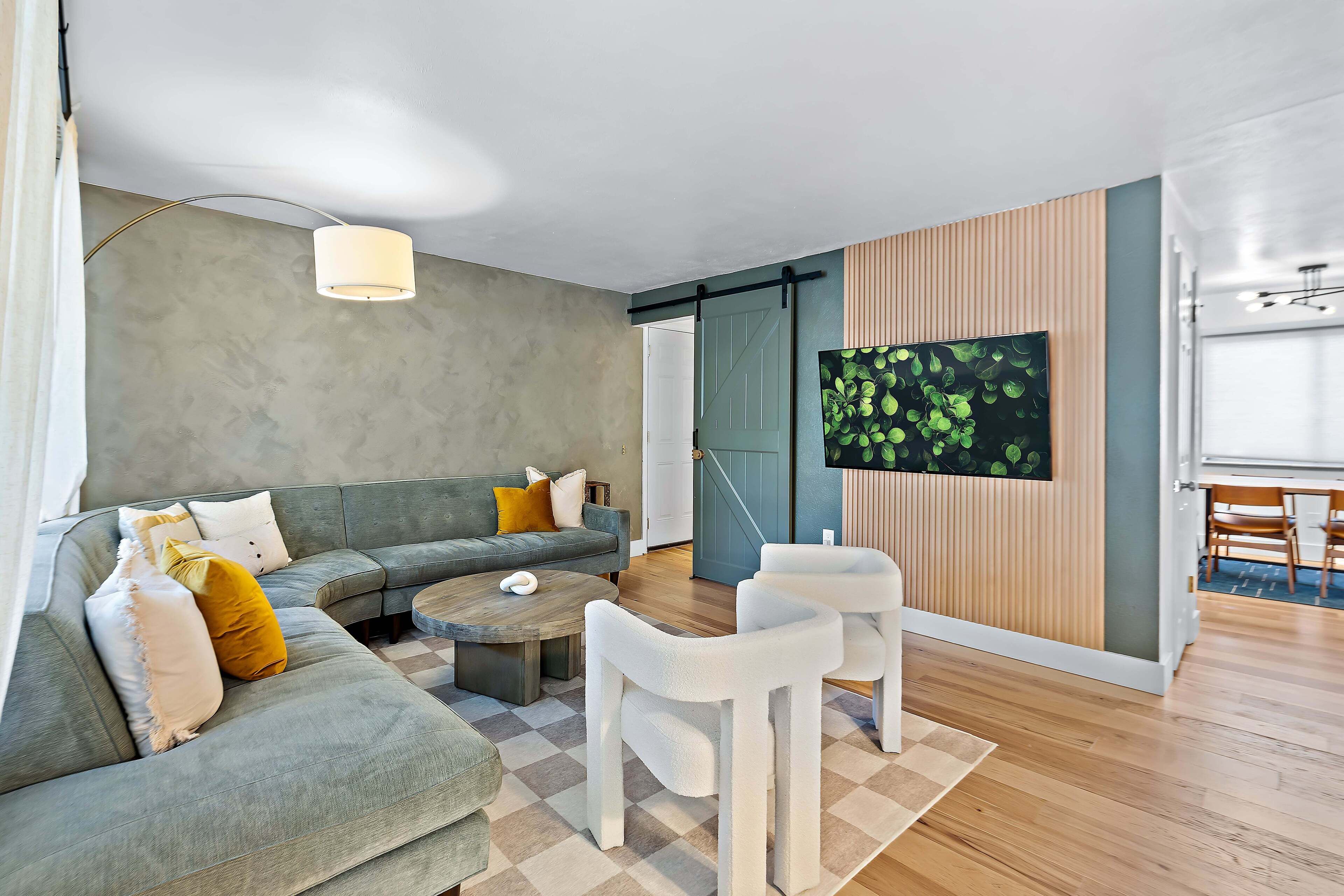 Un salon moderne avec un canapé gris, une table basse et des chaises blanches. Un téléviseur est fixé au mur et le sol est en bois.