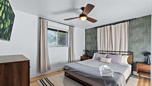 Un dormitorio moderno con cama, ventilador de techo, ventana con cortinas, mesita de noche y cómoda de madera.