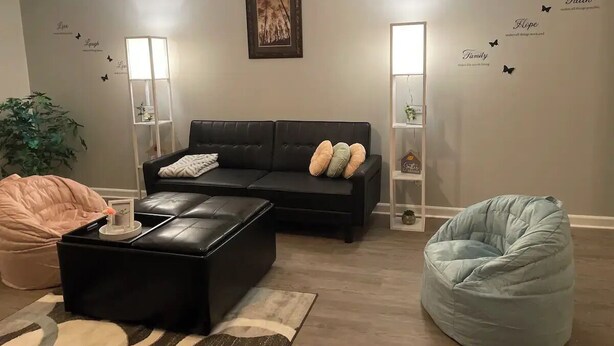 Ein modernes Wohnzimmer mit einer schwarzen Ledersofa, einem schwarzen Hocker, einem hellblauen Sitzsack und einem pinken Sitzsack. Zwei Stehlampen und ein gerahmtes Bild sind an der Wand zu sehen.