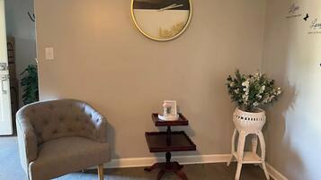 A room with a round mirror on the wall, a small wooden side table with a picture frame and a vase of flowers, a tufted armchair, and a wicker stool.