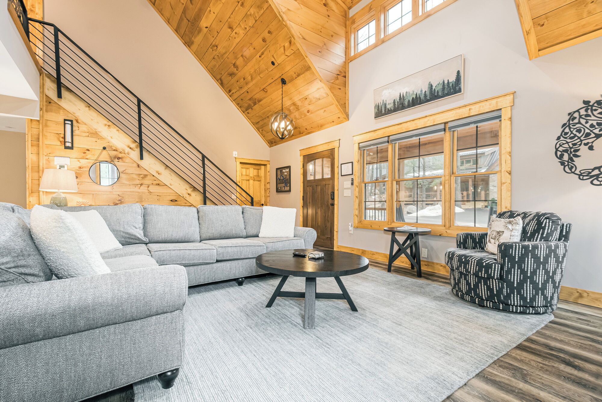 A spacious living room with a grey sectional sofa, a round coffee table, and a patterned armchair. A wooden staircase leads to an upper level.