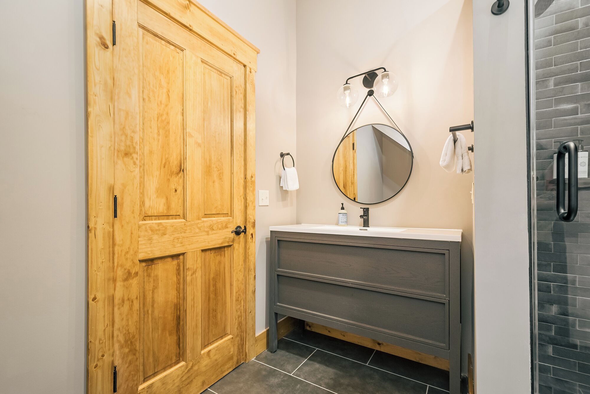 Un baño con puerta de madera, espejo redondo, lavabo y perchero para toallas.