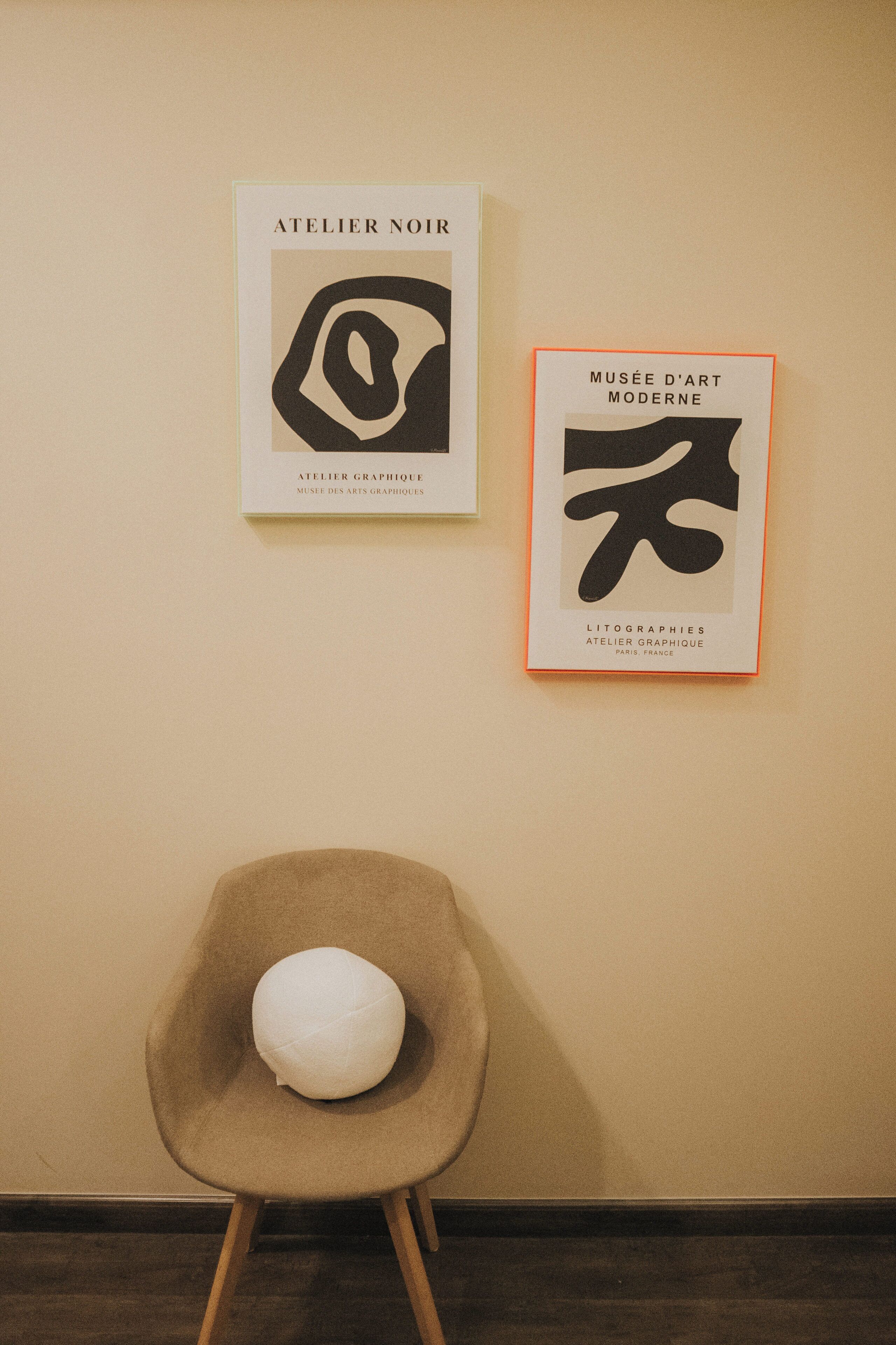 Twee ingelijste kunstwerken aan een muur, een stoel met een wit kussen ervoor.
