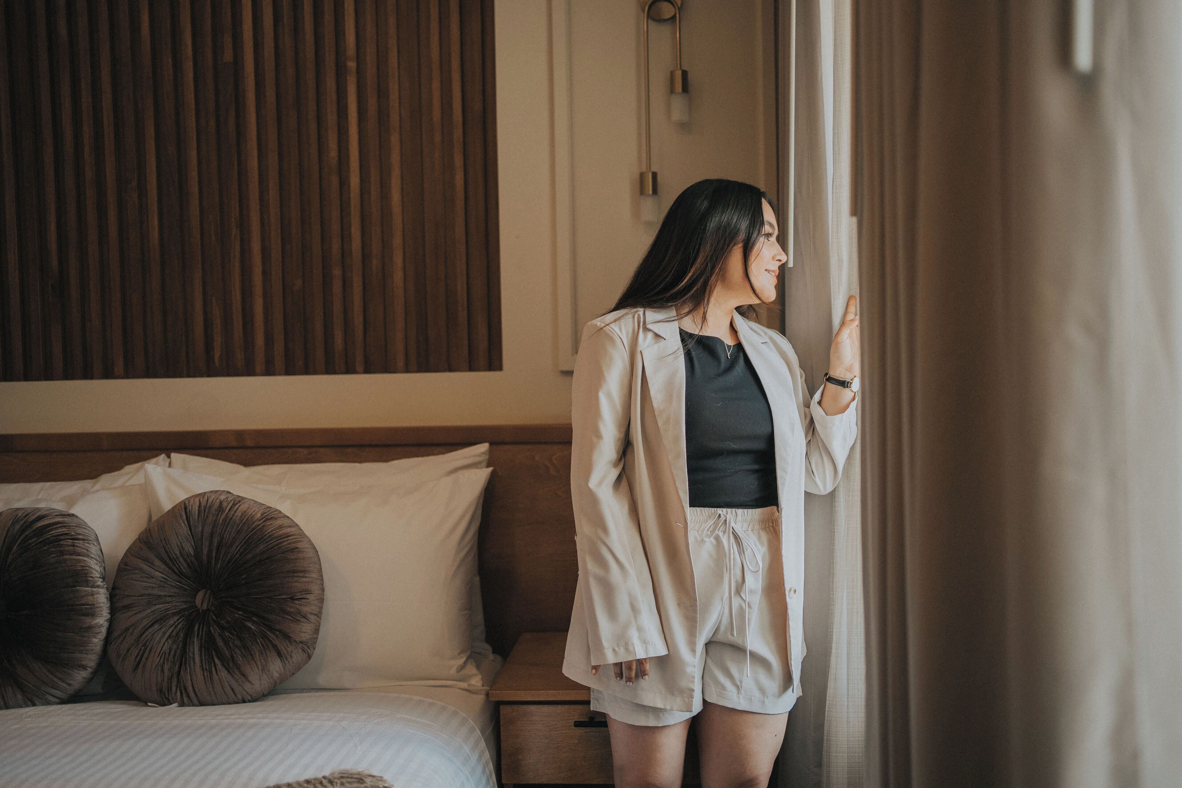 A person standing by a window in a bedroom with a wooden headboard, a bedside table, and two large round pillows.