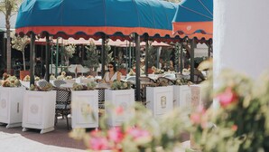 An outdoor seating area with tables and chairs under blue and red umbrellas, surrounded by potted plants and flowers. - Jules and Laurent Hotel Contemporaneo (Ensenada)