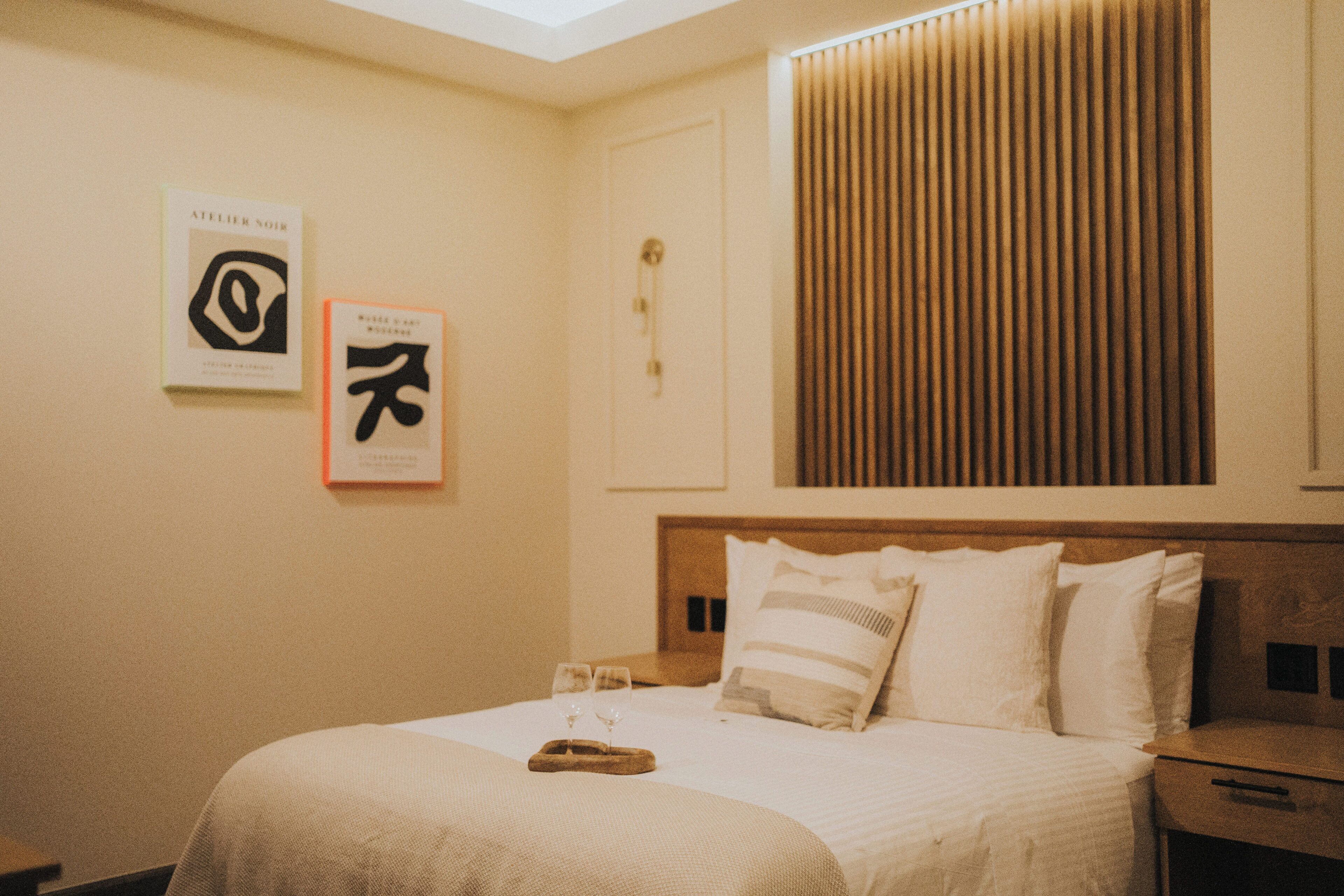 Habitación de hotel con una cama bien hecha, dos cuadros enmarcados en la pared, cabecera de madera y una ventana con persianas.