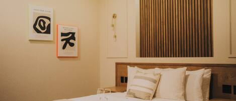 Habitación de hotel con una cama bien hecha, dos cuadros enmarcados en la pared, cabecera de madera y una ventana con persianas.