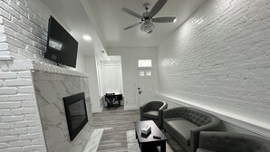 Un salon avec une cheminée, un téléviseur fixé au mur, un ventilateur de plafond et un mur en briques.