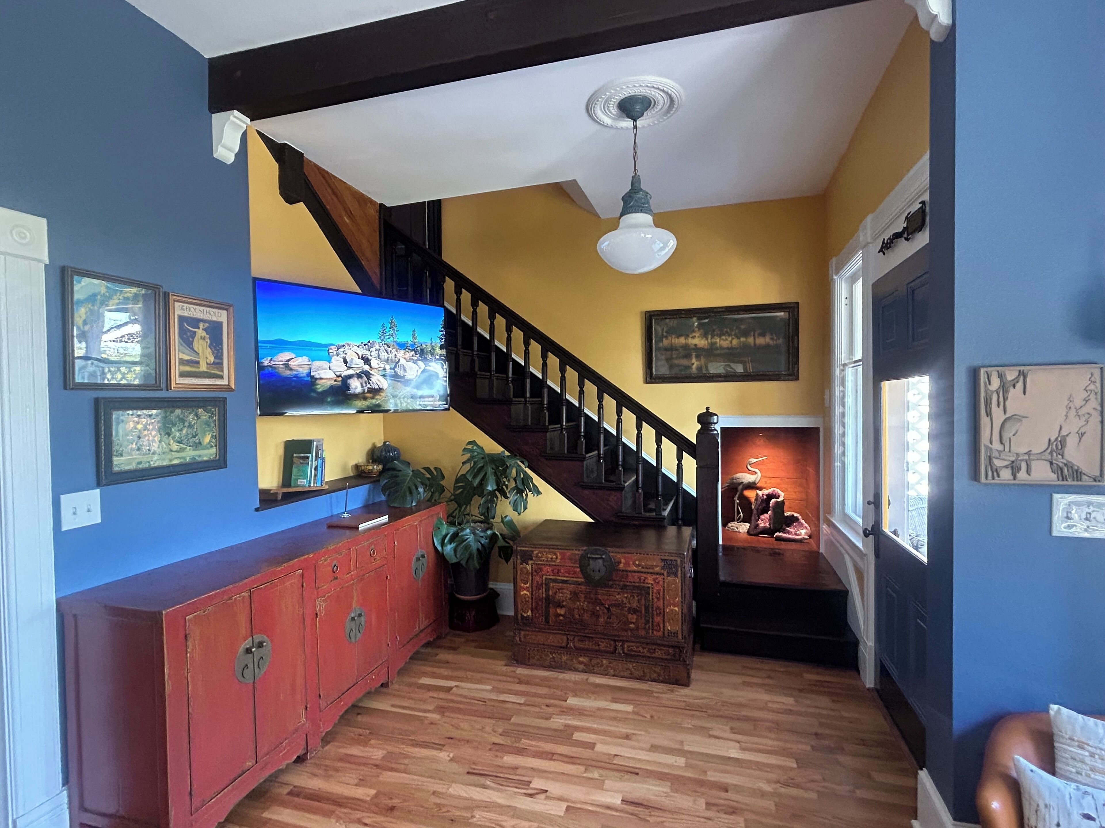 A room with a wooden floor, a red cabinet, a staircase, and a television mounted on the wall.