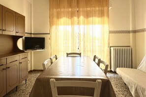 Een kamer met een eettafel, stoelen, een televisie op een dressoir, een radiator en een raam met gordijnen.