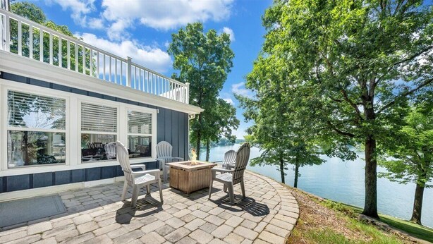 Un patio con mobiliario exterior y un hogar para fuego, con vista a un lago bordeado de árboles.